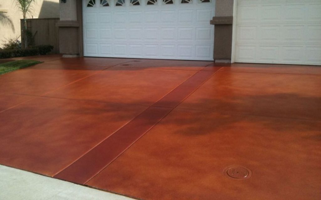 A wide driveway with a reddish-brown stained concrete finish, leading to a white garage door of a residential home. The smooth, uniformly colored surface is sectioned by control joints and features a manhole cover, highlighting a clean and well-maintained approach.