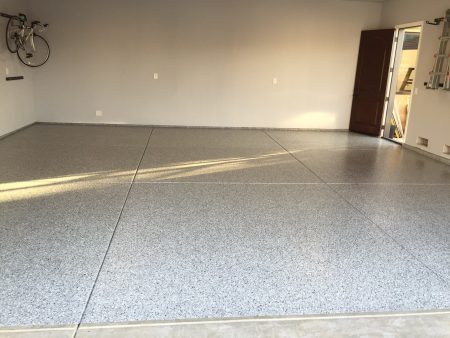 View from the driveway into a double garage with tan speckled epoxy flooring. The garage is open, revealing a clean, uncluttered space with two white doors on the back wall, a water heater to the right, and an electrical box on the left wall. The ceiling texture and walls are painted in a matching tan color, providing a uniform and tidy appearance.
