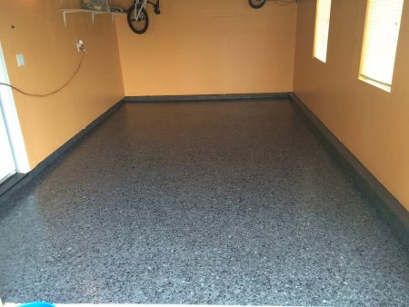 A room with a dark gray speckled epoxy floor and peach-colored walls. A bicycle hangs on a rack against the left wall, and two windows with blinds cast soft light into the room. An orange extension cord hangs from an outlet on the left. The room has a clean, organized appearance, likely serving as a storage or utility space.