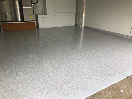 A room featuring a speckled gray epoxy floor with a shiny finish. To the left, there is a white water heater and a set of white storage cabinets above an exposed wooden base. A closed white door is on the right, and in the foreground, a white boundary line marks the edge of the flooring area. The space appears clean and is likely part of a residential garage or utility area.