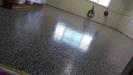 Interior of a room with glossy, speckled epoxy flooring reflecting natural light from windows. The space includes a beige wall with two coiled black cables hung neatly on the right. In the back left corner, a wooden pallet with items wrapped in clear plastic is partially visible. The area conveys a tidy, utilitarian atmosphere with ample lighting.
