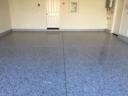 A clean, empty room with a light gray speckled epoxy floor. There are two white doors on the back wall, one closed and the other slightly open. To the right, there's a water heater and gas pipeline. The room also features a small white electrical panel on the right wall. The space looks like a functional area, possibly a laundry or utility room in a residential home.