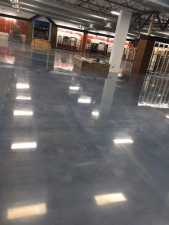 An expansive, well-lit interior space with a high ceiling and reflective polished concrete floor. The area has a variety of fixtures, including a reception desk with a stone facade, red and wood-toned wall accents, and metal-framed glass partitions. Ceiling-mounted lights cast a series of bright reflections on the floor. It looks like a commercial setting, possibly a lobby or retail environment, with some areas cordoned off, indicating potential construction or rearrangement.