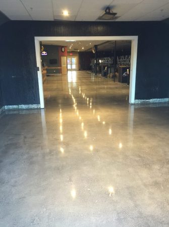 A view from a darkened area into a well-lit, spacious room with a reflective concrete floor. The room features a bar area with stools and a television displaying a sports channel. On the right, an open doorway leads to a further brightly colored space. The shiny floor surface captures the ceiling lights, creating a pattern of bright spots that lead the eye into the room. The setting suggests a social venue, like a pub or restaurant, preparing to welcome patrons.
