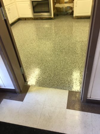 ALT Text: "View from a doorway looking into a kitchen with a multi-colored speckled epoxy floor. The transition from a beige tiled area to the epoxy floor is visible, leading to white kitchen cabinetry with some areas exposed for repair. The space appears to be in the process of home improvement or renovation.