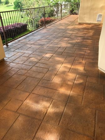 Covered patio with terracotta-colored stamped concrete flooring, leading to an ornate wrought iron railing overlooking a lush green landscape, invoking a sense of warmth and serene outdoor living.