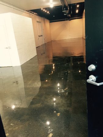 View from behind a door partially ajar, showing a large room with a wet concrete floor reflecting the overhead lights, with white painted brick walls and a sense of depth created by the lighting.