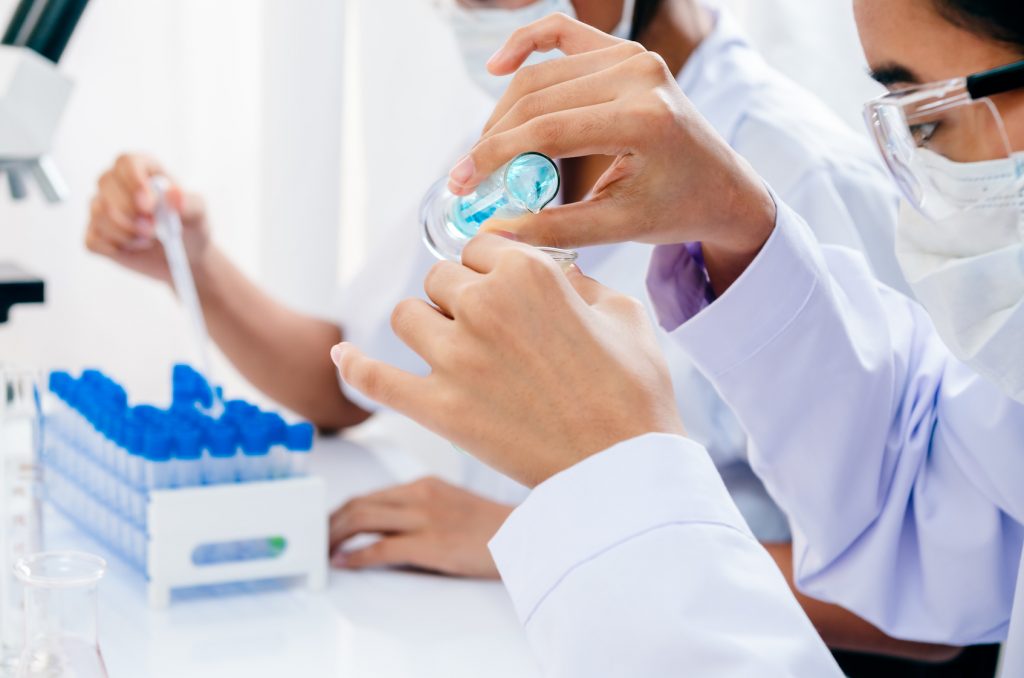 Close-up of two scientists in a laboratory, one handling a pipette and a test tube with a blue liquid. Both are wearing white lab coats, protective gloves, and safety glasses, adhering to safety protocols. The focus on their hands and equipment underscores the precision and concentration required in scientific research.