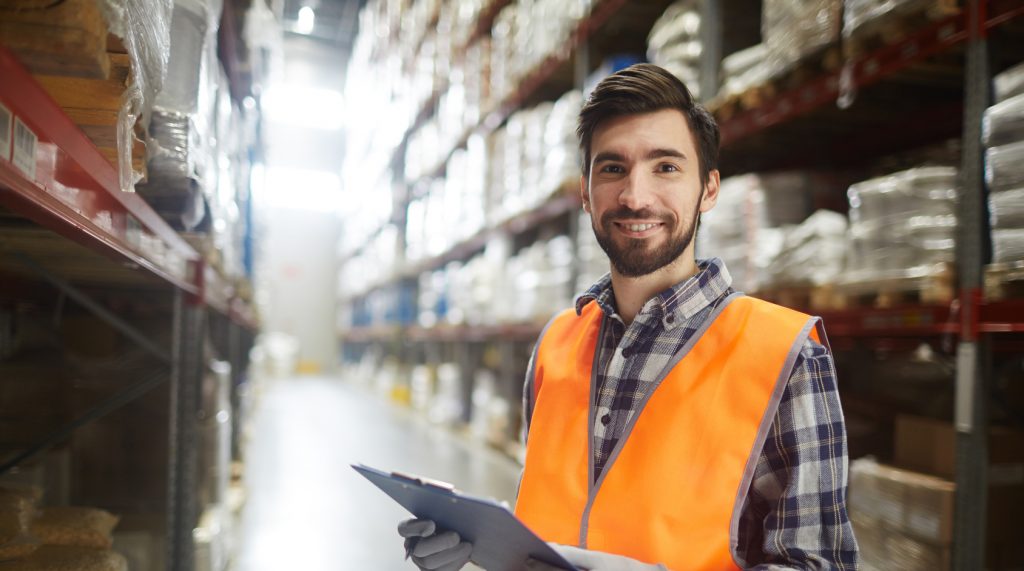 A confident male warehouse worker in a high-visibility orange vest stands in an aisle between tall shelves stocked with goods. He holds a clipboard, possibly taking inventory or managing orders. The large warehouse setting indicates an industrial or commercial environment, and the worker's pleasant expression suggests a positive workplace.
