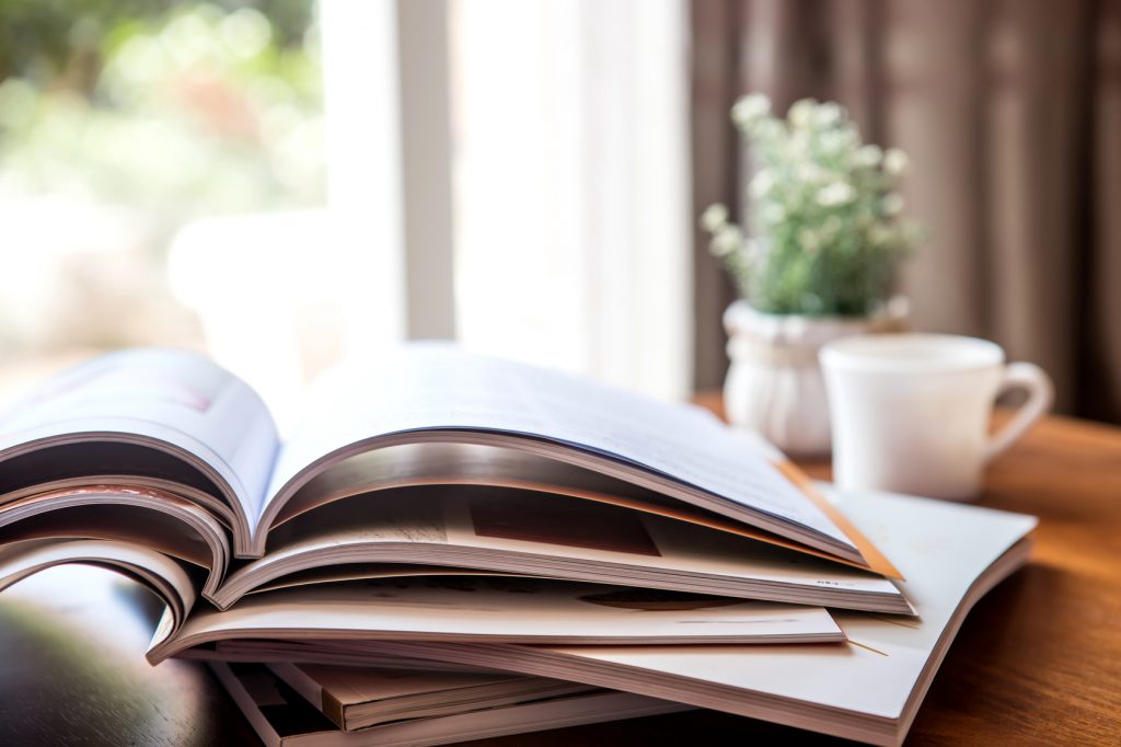 An open book with glossy pages lies on a wooden table, with a blurred background featuring a small potted plant and a white coffee cup. The cozy setting suggests a peaceful reading environment, possibly in a home or a café, with natural light streaming in from a nearby window.