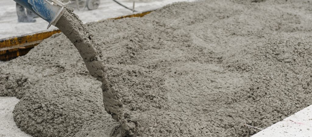 A stream of wet concrete flowing from a chute to form a new slab. The image captures the concrete in motion, displaying its thick texture as it spreads over the area with reinforcing steel bars partially visible, indicating ongoing construction work.