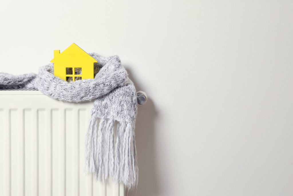 A simplistic yellow paper house perched on a white radiator, wrapped in a cozy grey knitted scarf with tassels. The scene evokes a sense of warmth and home comfort, suggesting energy-saving or staying warm during winter.