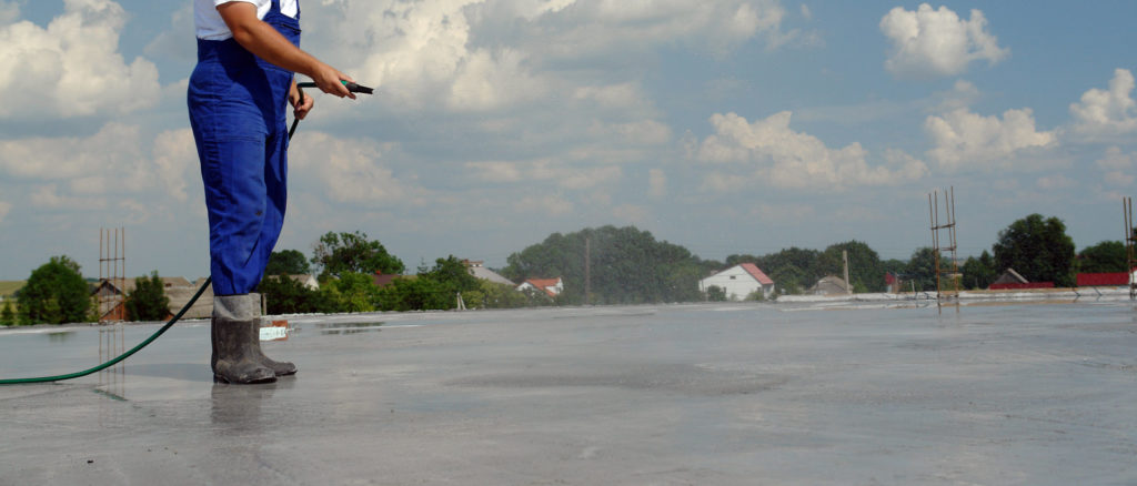 A worker in a blue uniform and rubber boots using a hose to clean a large concrete surface on a rooftop, with a village and cloudy sky in the background.