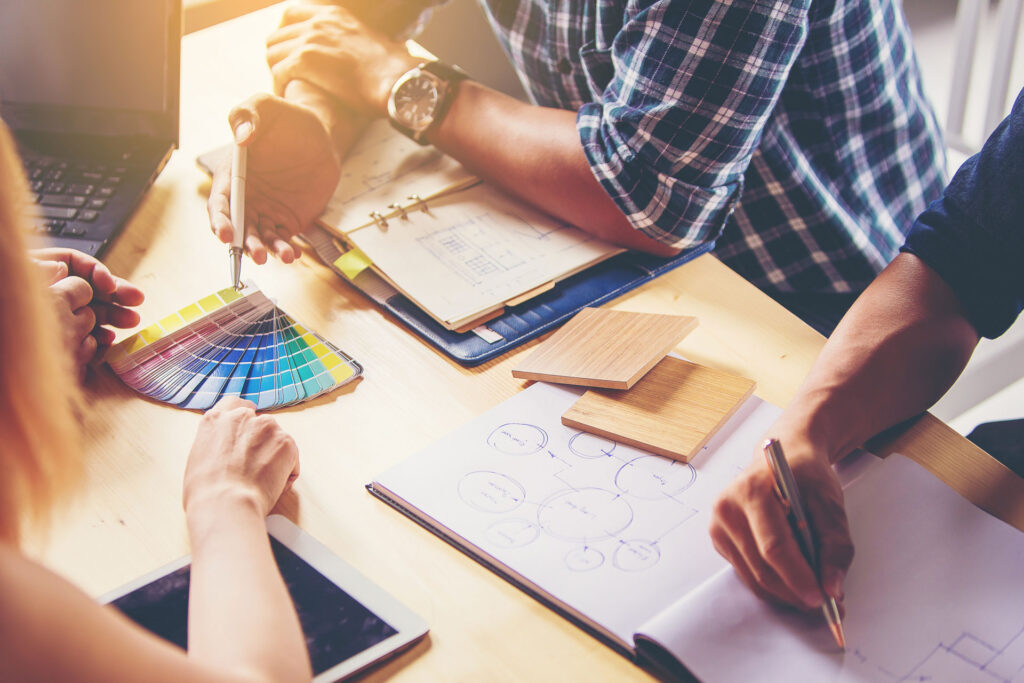 A team collaborating around a table with design tools: a person holding a color swatch, another drawing a flowchart, and samples of wood veneer alongside a laptop and tablet, indicative of a creative brainstorming session.
