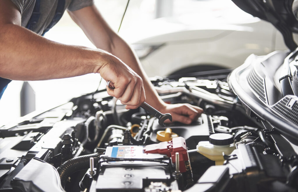 A mechanic's hands using a wrench on a car engine, showcasing a close-up of automotive maintenance work with various engine components and a car battery in the foreground.