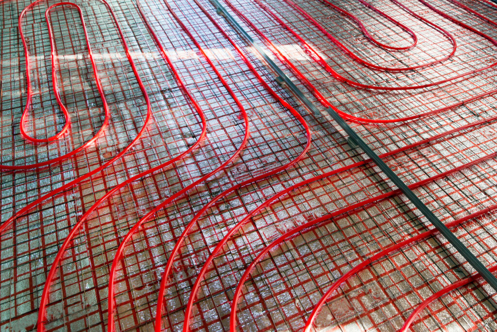Close-up of a red underfloor heating installation with winding pipes laid out on a mesh support over a concrete substrate, illustrating the process of installing a radiant floor heating system.