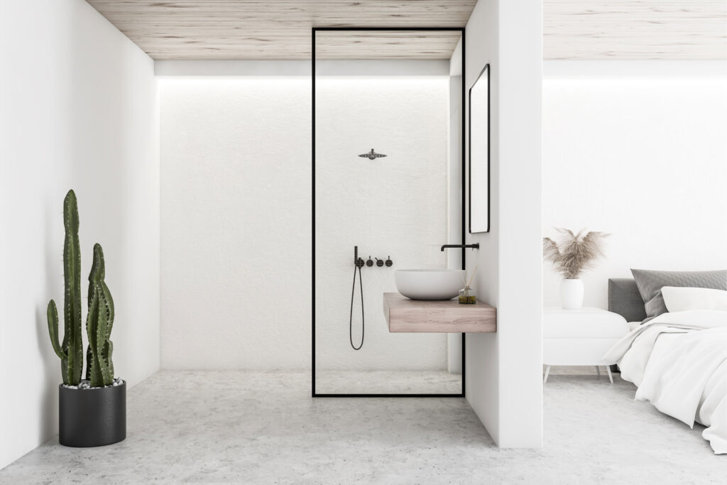 Minimalist interior design of a bedroom with an adjoining open bathroom. The bedroom features a low-profile bed with soft linens, while the bathroom includes a modern basin on a wooden shelf, black fixtures, and a glass shower partition. A tall cactus plant adds a touch of greenery to the neutral color scheme.