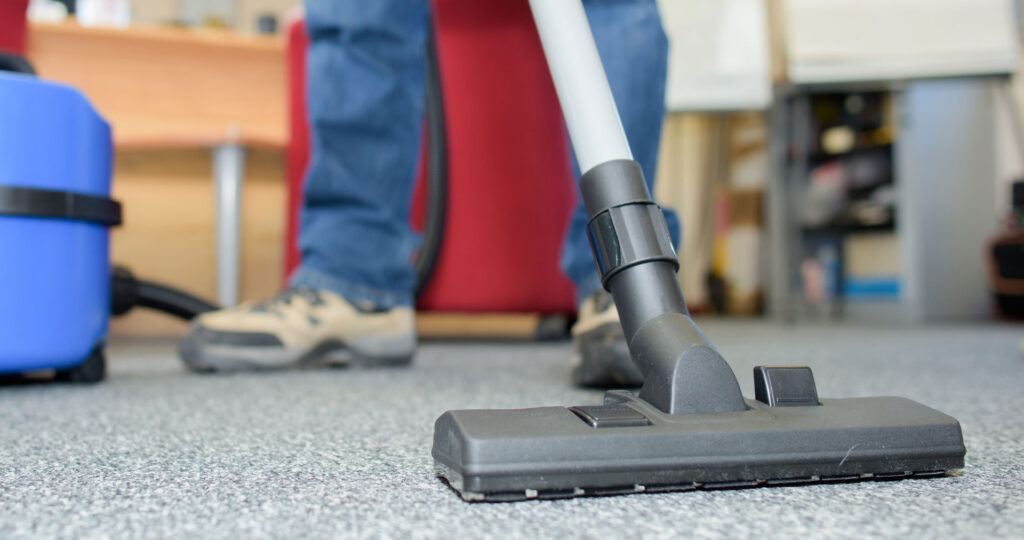 A close-up view of a vacuum cleaner's nozzle on a textured office carpet, with the legs of a person operating it in the background, implying routine cleaning or maintenance work being done in an office environment.