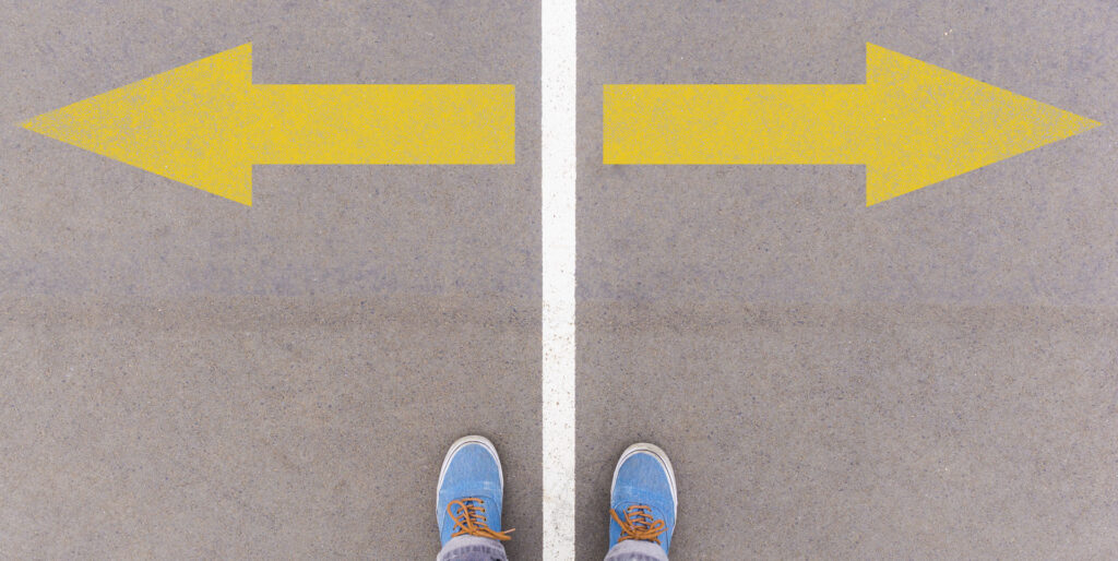 First-person view of a person's feet standing on a gray surface with two large yellow arrows pointing in opposite directions, symbolizing a crossroad or decision-making point.