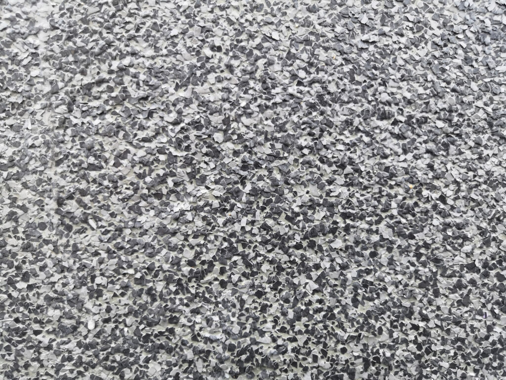 Close-up of a rough-textured surface with a speckled pattern of small, irregular black, gray, and white flakes, resembling a pebble dash or terrazzo floor finish.