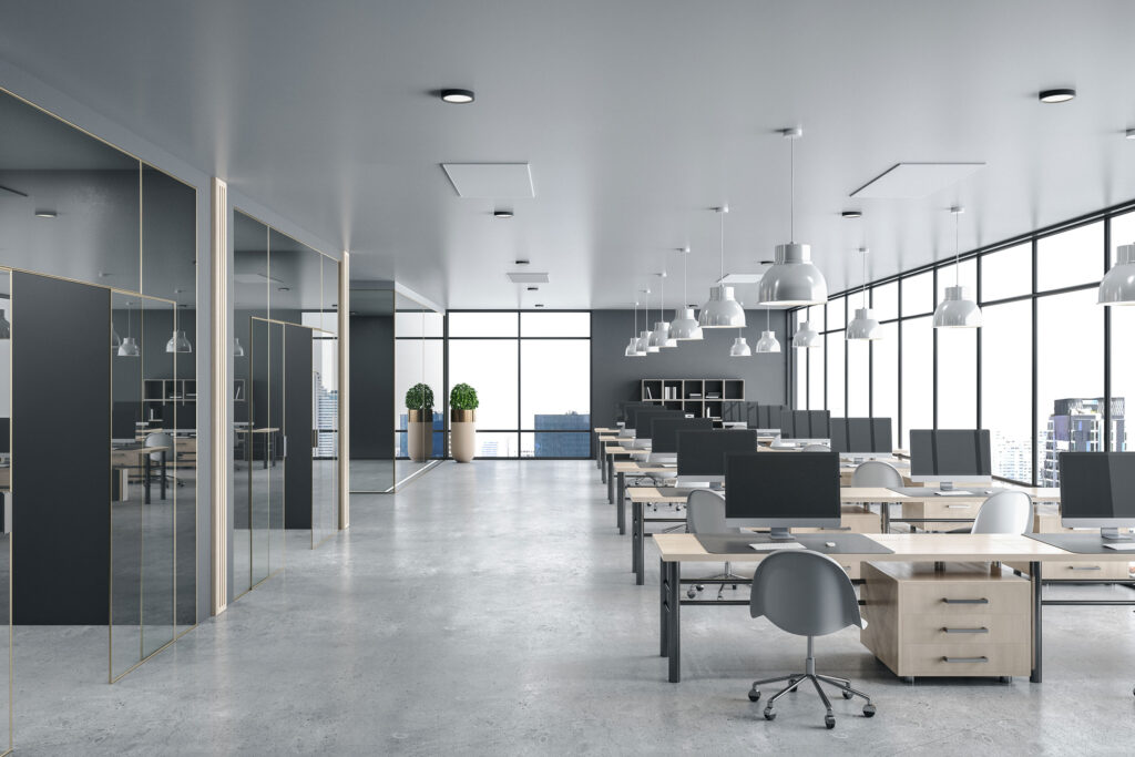 Modern open-plan office with rows of wooden desks equipped with desktop computers and gray ergonomic chairs on a polished concrete floor. Large floor-to-ceiling windows line the right wall, offering views of city skyscrapers. The ceiling features multiple white pendant lights, and two potted plants stand near the windows at the back. Glass-walled private offices or meeting rooms are on the left side. The space has a sleek, minimalistic design with neutral gray and natural wood tones.